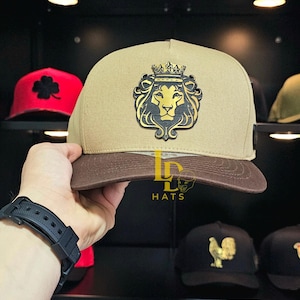 May include: Tan and brown baseball cap featuring a gold and black lion crest with a crown. The cap has a flat brim and is held up by a person. The text "LD HATS" is visible on the cap.