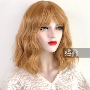 May include: A light brown wavy bob wig with bangs. The wig is styled with loose waves and a side part. The bangs are cut to just above the eyebrows.