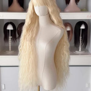 May include: A long, wavy blonde wig with a blunt bang is displayed on a mannequin. The wig is a light, creamy color and has a natural-looking texture. Other wigs in various styles and colors are visible in the background.