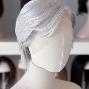 以下が含まれることがあります： マネキンの頭に展示された、サイドパートのある短いシルバーグレーのウィッグ。ウィッグは自然なヘアラインで、おそらく合成繊維でできています。背景には他のウィッグが見えます。