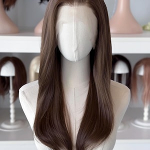 Puede incluir: Una peluca larga y lisa de color marrón medio con raya en medio y frente de encaje transparente. El cabello cae por debajo de los hombros con una textura suave y natural. La peluca se muestra sobre un maniquí de color neutro.