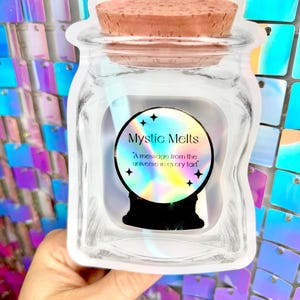 K&ouml;nnte beinhalten: Ein klares Glas mit einem Korkdeckel, das ein Etikett mit dem Text "Mystic Melts" und "A message from the universe in every tart" zeigt. Das Etikett zeigt ein Kristallkugel-Design mit Sternen.