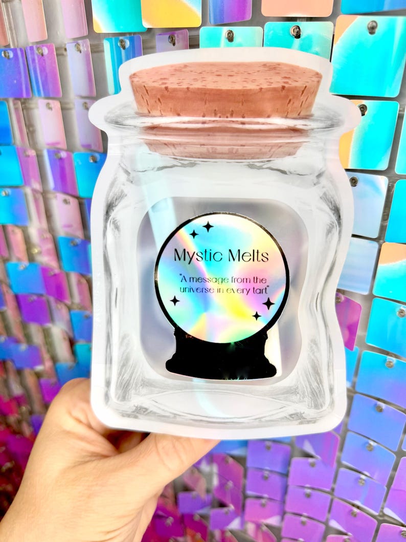 K&ouml;nnte beinhalten: Ein klares Glas mit einem Korkdeckel, das ein Etikett mit dem Text "Mystic Melts" und "A message from the universe in every tart" zeigt. Das Etikett zeigt ein Kristallkugel-Design mit Sternen.