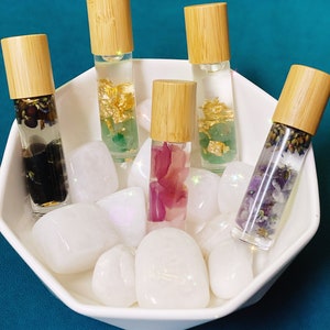 May include: Five glass roller bottles with wooden caps filled with different crystals and flowers. The bottles are sitting in a white bowl with white stones.
