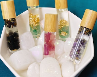 May include: Five glass roller bottles with wooden caps filled with different crystals and flowers. The bottles are sitting in a white bowl with white stones.