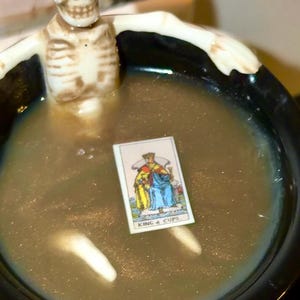 K&ouml;nnte beinhalten: Ein schwarzer Kessel gef&uuml;llt mit einer schimmernden, goldenen Fl&uuml;ssigkeit, mit einer kleinen Skelettfigur und einer Tarotkarte mit der Aufschrift "KING 4 CUPS". Der Kessel hat wei&szlig;e Schrift auf der Seite. Eine einzigartige Halloween-Dekoration.