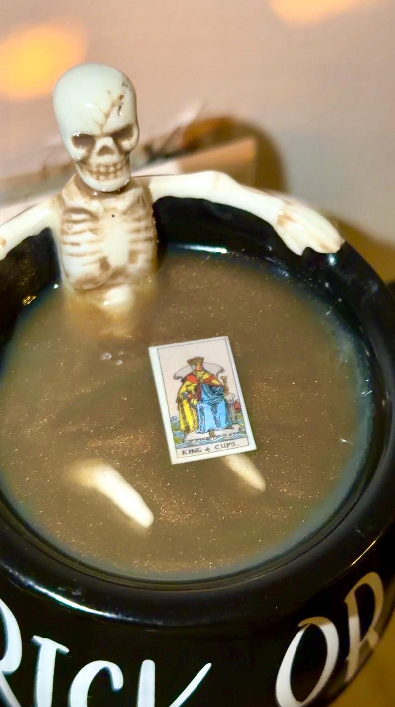 K&ouml;nnte beinhalten: Ein schwarzer Kessel gef&uuml;llt mit einer schimmernden, goldenen Fl&uuml;ssigkeit, mit einer kleinen Skelettfigur und einer Tarotkarte mit der Aufschrift "KING 4 CUPS". Der Kessel hat wei&szlig;e Schrift auf der Seite. Eine einzigartige Halloween-Dekoration.