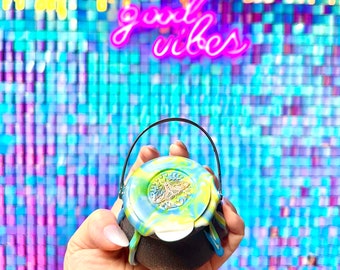 May include: A small black cauldron with a green, blue, and white swirl design. The cauldron has a handle and a gold butterfly design on the lid. The cauldron is being held in a hand against a background of iridescent blue and purple squares with the words "good vibes" in pink neon.