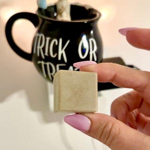 K&ouml;nnte beinhalten: Ein kleiner, quadratischer, hellfarbiger Gegenstand wird vor einer schwarzen Tasse mit den Worten "TRICK OR TREAT" in wei&szlig;er Schrift gehalten. Die Tasse hat einen Henkel und ist mit einer kleinen Skelettfigur verziert.