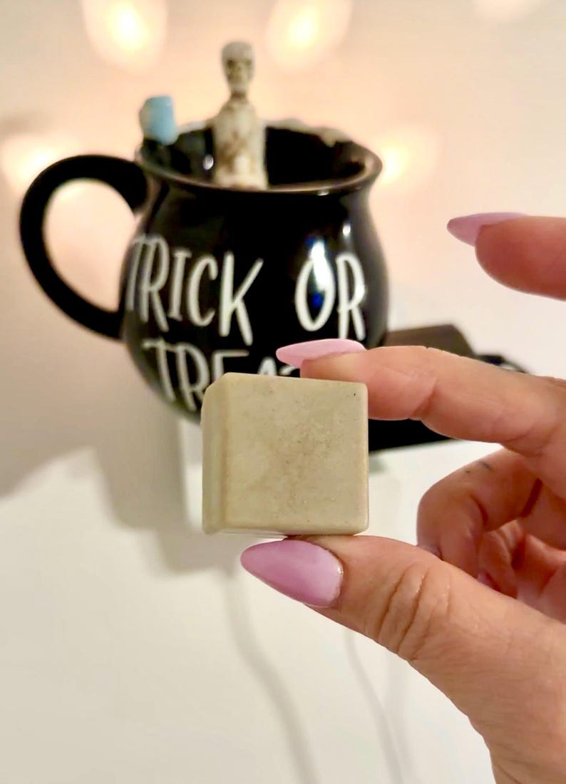 K&ouml;nnte beinhalten: Ein kleiner, quadratischer, hellfarbiger Gegenstand wird vor einer schwarzen Tasse mit den Worten "TRICK OR TREAT" in wei&szlig;er Schrift gehalten. Die Tasse hat einen Henkel und ist mit einer kleinen Skelettfigur verziert.