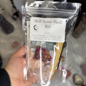 May include: A clear plastic bag labeled "Self-Love Spell Kit" containing a spell jar, rose quartz chips, incense, Palo Santo, pink Himalayan salt, sugar, rose petals, lavender, pink wax, and a melting spoon. The kit is held in a hand.