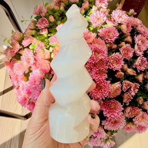 Puede incluir: Una escultura en espiral de selenita blanca se sostiene frente a un ramo de flores rosas. La escultura tiene un diseño en capas y retorcido. Las flores son de un color rosa vibrante y el fondo está borroso.