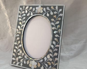 Op de afbeelding: Een grijs fotolijstje met een ovale opening, versierd met een bloemmotief van parelmoer.