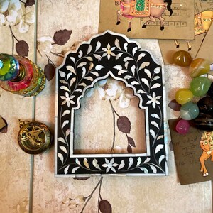 May include: A black and white decorative frame with floral and leaf inlay. The frame has an arched top. Other decorative items surround the frame, including a sundial, perfume bottle, and gemstones.