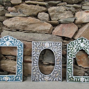 Op de afbeelding: Drie decoratieve fotolijsten met bloemen- en bladerontwerpen. De lijsten zijn in teal, grijs en groen, met parelmoer accenten. De lijsten hebben verschillende boog- en ovale openingen.
