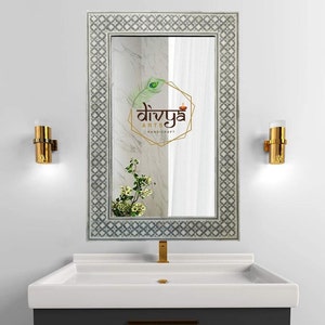 Puede incluir: Un tocador de baño blanco con un lavabo de porcelana blanca y un grifo dorado. Un espejo rectangular grande con un marco de mosaico gris y blanco cuelga sobre el lavabo. Dos apliques de pared dorados están a cada lado del espejo. El espejo refleja una pared de mármol blanco con un logotipo que dice "Divya Arts Handicraft".