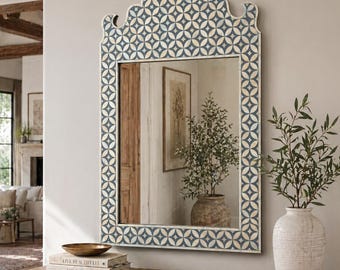 May include: A rectangular wall mirror with a decorative, arched top. The frame features a repeating pattern of blue and white geometric shapes. The mirror is displayed above a wooden dresser, with a vase of greenery to the right.