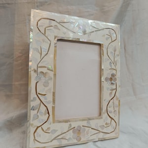 Puede incluir: Un marco de fotos blanco con un diseño floral hecho de nácar. El marco tiene una forma rectangular y un fondo blanco.