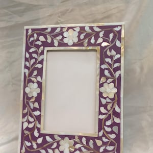 Könnte beinhalten: Ein lilafarbener Bilderrahmen mit einem weißen floralen Design aus Perlmutt. Der Rahmen ist rechteckig und mit filigranen Details verziert.