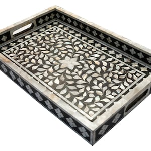 May include: A black rectangular tray with a white mother of pearl inlay design. The tray has a floral pattern with leaves and a flower in the center. The tray has a decorative border with a geometric pattern.