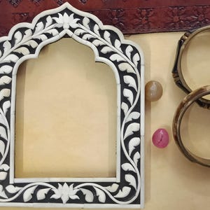 May include: A white and black ornate picture frame with a floral design. The frame has an arched top and is made of bone inlay. Two gold bangles are on the right side of the frame.