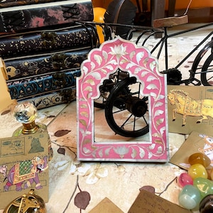 May include: A pink and white decorative frame with floral designs and a carved archway. The frame is made of mother of pearl and has a pink background. The frame is sitting on a table with other decorative items.