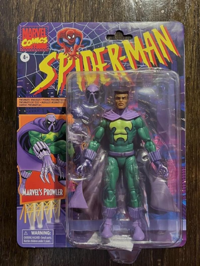 May include: A Marvel's Prowler action figure in a plastic package. The figure is green and purple with a black mask. The figure is holding its hands up in a fighting pose. The package has a picture of Spider-Man on it.