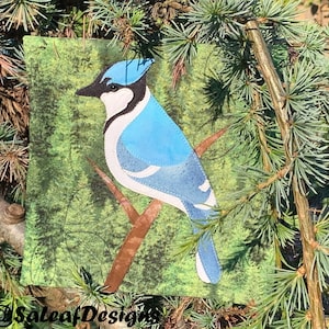 May include: A blue jay with a white chest and blue wings perched on a branch in a green forest setting. The bird is made of fabric and is a quilted wall hanging.