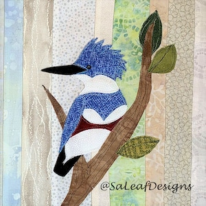 May include: A blue and white bird with a red breast perched on a brown branch with green leaves. The bird is a kingfisher and is sitting on a branch with green leaves. The background is a patchwork of different fabrics.