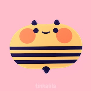 May include: A yellow and black bee-shaped mousepad with a smiling face and orange cheeks. The mousepad has a pink background and the text "tinkalila" at the bottom.
