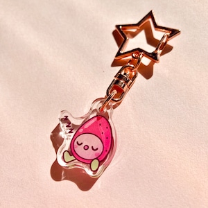 May include: A pink and green cartoon strawberry with a sleepy face is printed on a clear acrylic keychain. The keychain is attached to a gold star-shaped metal ring.