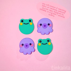 Pode incluir: Quatro tampas de Joy-Con de silicone coloridas, duas com um design de sapo verde com olhos amarelos e duas com um design de polvo roxo com bochechas rosadas. O texto "you will receive 2 matching pairs of randomly selected joy-con caps designed by tinkalila, in b-grade / seconds quality" &eacute; vis&iacute;vel no canto superior direito da imagem. O texto "tinkalila" &eacute; vis&iacute;vel no canto inferior direito da imagem.