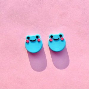 May include: Two blue frog-shaped charms with pink cheeks and black eyes. The charms are on a pink background.