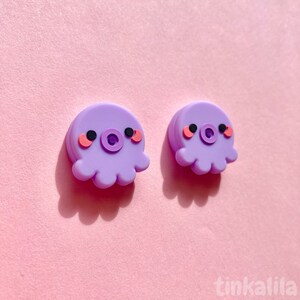 Puede incluir: Un par de pendientes de pulpo morado con mejillas rosadas y ojos negros. El pulpo tiene una peque&ntilde;a boca redonda.
