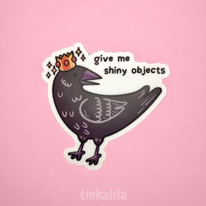 Puede incluir: Una pegatina con un cuervo de dibujos animados que lleva una corona dorada y el texto "give me shiny objects". El cuervo es negro con un pico y patas moradas. La pegatina tiene un borde blanco y está sobre un fondo rosa.