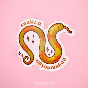 Puede incluir: Una serpiente amarilla y naranja con estrellas a su alrededor. La serpiente está enroscada y el texto "snake it till you make it" está escrito a su alrededor.