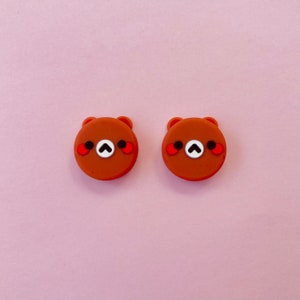 May include: Two brown bear-shaped silicone buttons with red cheeks and black eyes. The bears have a white nose and a white heart shape on their chest.