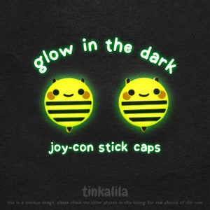 Puede incluir: Dos tapas para joystick Joy-Con en forma de abeja, de color amarillo y negro, que brillan en la oscuridad. El texto "glow in the dark" y "joy-con stick caps" está escrito en verde.