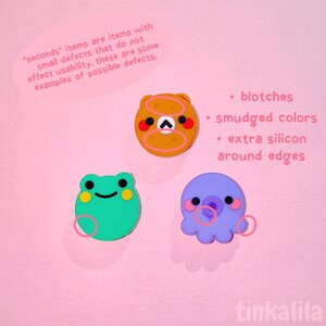 Pode incluir: Tr&ecirc;s agarradores de telefone de silicone em forma de sapo, polvo e urso. O sapo &eacute; verde com olhos amarelos, o polvo &eacute; roxo com a boca rosa e o urso &eacute; castanho com as bochechas laranja. O texto "seconds" items are items with small defects that do not effect usability. these are some examples of possible defects. &bull; blotches &bull; smudged colours &bull; extra silicon around edges" est&aacute; escrito em branco sobre um fundo rosa.