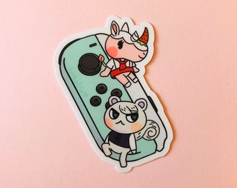 Puede incluir: Un mando de juego Nintendo Switch Joy-Con verde y blanco con dos personajes de animales en la parte superior. El personaje de la izquierda es una ardilla blanca con ojos negros y un chaleco negro. El personaje de la derecha es una oveja blanca con mejillas rosadas y un vestido rojo. El texto "Tinkalila" está escrito en el mando.