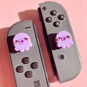 Pode incluir: Dois controladores cinzentos do Nintendo Switch com capas de bot&otilde;es em forma de polvo roxo. Os controladores t&ecirc;m os bot&otilde;es padr&atilde;o: X, Y, A, B e um bot&atilde;o home.