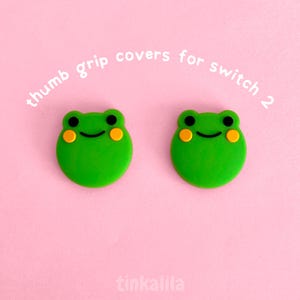 Può includere: Due protezioni per impugnatura del pollice a forma di rana verde per Switch 2, con occhi neri e guance gialle, su sfondo rosa. Il testo "thumb grip covers for switch 2" è arcuato sopra. Il marchio "tinkalila" è in basso.