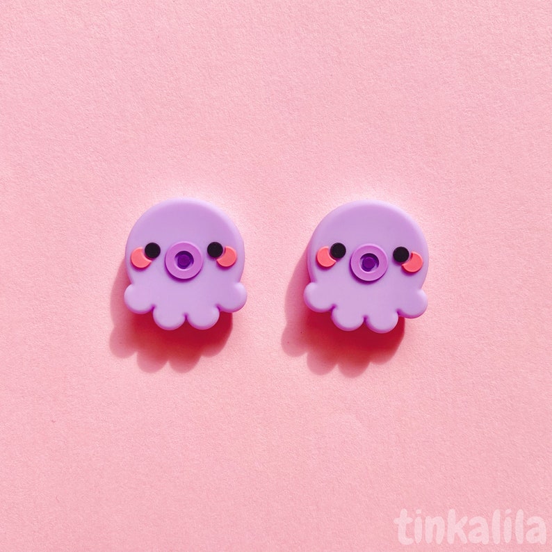 Puede incluir: Dos pendientes de color morado en forma de pulpo con mejillas rosadas y ojos negros. Los pendientes est&aacute;n sobre un fondo rosa.