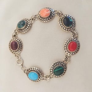 May include: Silver bracelet featuring seven oval gemstones in shades of red, green, blue, and orange. Each stone is set in a decorative silver bezel and linked together with a chain. The bracelet is designed for wrist wear.