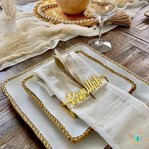 May include: Elegant table setting featuring white square plates with gold beaded rims, a folded white linen napkin, and a gold napkin ring with script. The table is set on a wooden surface, with a decorative runner and glassware.