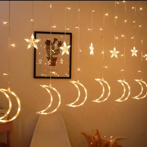 May include: String lights with warm white LED lights shaped like stars and crescent moons. The lights hang from a thin wire, creating a celestial display. The lights are arranged in a vertical pattern, with a picture in the background.