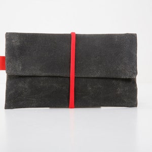 Puede incluir: Cartera de lona gris oscuro con cierre de banda elástica roja. La cartera tiene un cierre de solapa y una pequeña pestaña roja en el lateral. La lona tiene un aspecto ligeramente desgastado, lo que sugiere un estilo vintage o robusto.