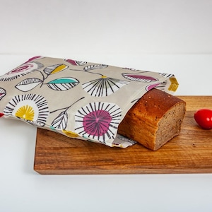 Könnte beinhalten: Ein Brotlaib teilweise in einer Brotbeutel mit floralem Muster, auf einem Holzschneidebrett. Eine kleine rote Tomate liegt neben dem Brot. Die Tasche hat einen beigen Hintergrund mit bunten floralen Designs.