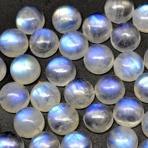 May include: A collection of round, white moonstone cabochons with iridescent blue flashes. The stones are smooth and polished, and they are arranged in a random pattern.