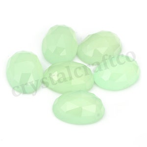 May include: Six pale green oval-shaped gemstones with faceted edges. The gemstones are arranged in a cluster on a white surface.